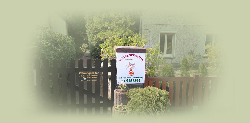 Holzzaun mit offenem Tor, an dem ein Schild mit der Aufschrift 'Katzenpension' und einer roten Katzenzeichnung hängt, davor ein kleiner Busch und Blumen.